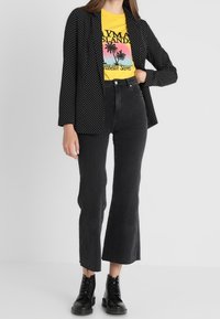 Czarny żakiet w kropki noszony na żółtej koszulce z grafiką, w połączeniu z czarnymi, wysokim pasem dżinsami z rozszerzanymi nogawkami oraz czarnymi botkami na sznurówki.