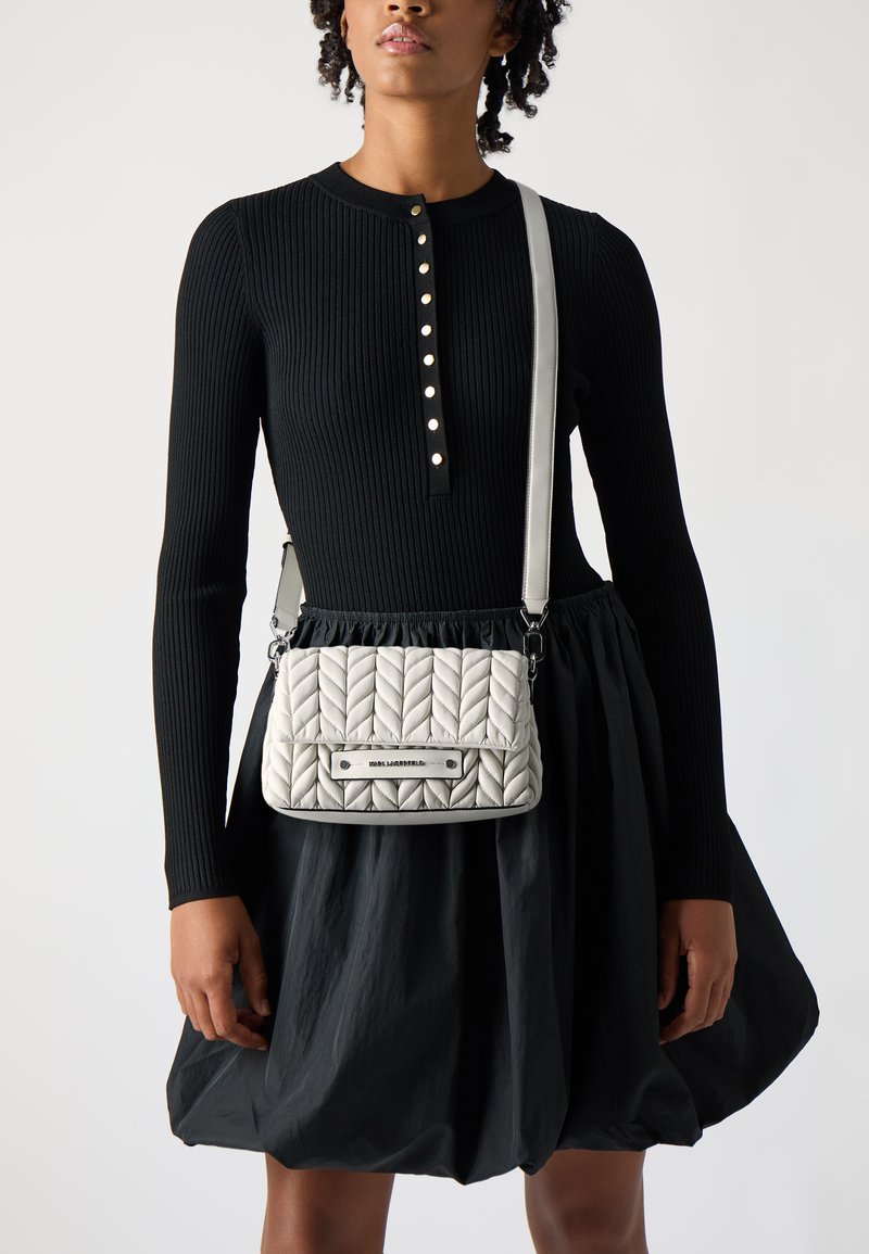 Borsa a tracolla grigia trapuntata con un motivo intrecciato decorativo, hardware argentato e una tracolla rimovibile, abbinata a un vestito nero a coste.