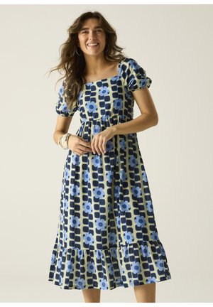 Donna sorridente con un vestito midi a fantasia floreale blu e nero, maniche a sbuffo e orlo arricciato, in piedi davanti a uno sfondo semplice.