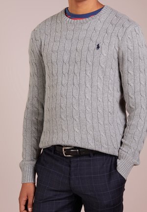 Mężczyzna ubrany w szary sweter warkoczowy na niebieską koszulę i ciemnoniebieskie spodnie w kratę z czarnym paskiem, ręka w kieszeni.