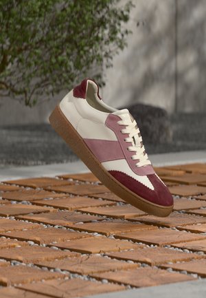 Hvid lædersneaker med bordeaux og lyserøde ruskindsdetaljer, med en tekstureret sål og klassisk snøredesign, hævet over en træoverflade.