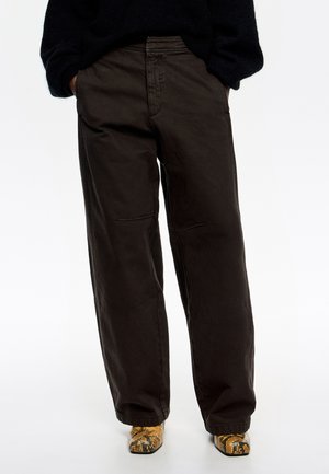 Szerokie, ciemnobrązowe spodnie bawełniane o luźnym kroju, z delikatnymi detalami w szwie, połączone z obuwiem w żółto-czarny wzór.