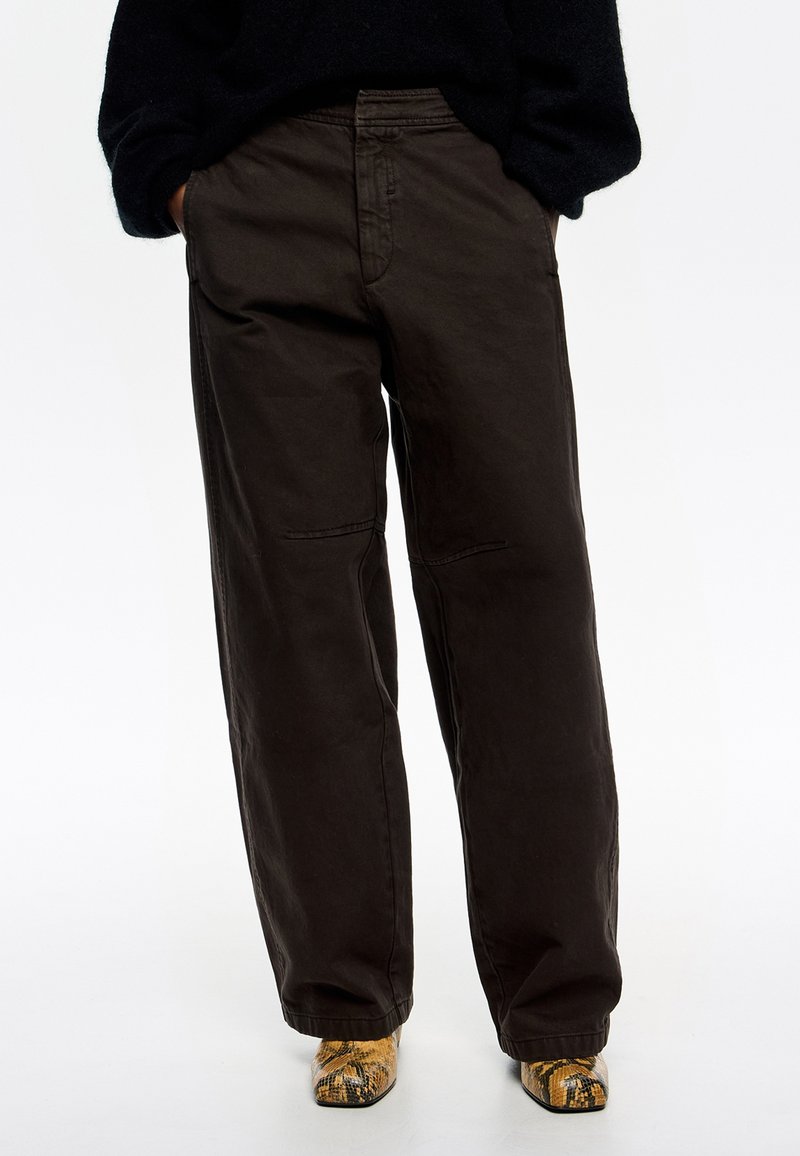 Pantalones de algodón oscuros de corte amplio y ajuste relajado, con detalles de costura sutiles, combinados con calzado amarillo y negro de estampado.