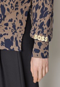 Manche d'un pull marron et bleu foncé à motifs avec "BOSS" brodé près du poignet au-dessus de la main d'une personne, devant un pantalon noir.