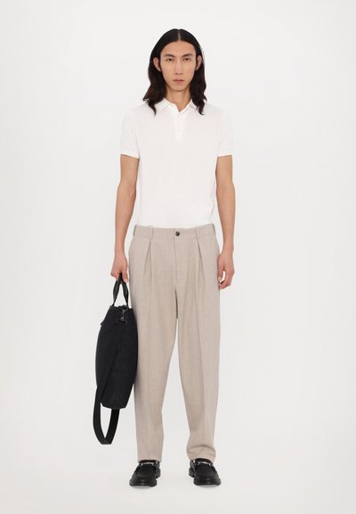 Chemise polo blanche, pantalon beige plissé, sac noir. Le modèle se tient droit avec des cheveux à longueur d'épaule et des chaussures noires. Fond simple.
