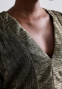 Metallisch gouden textielstof met verticale lijnen, met een V-halsontwerp en elegante draping. Korte, close-up van het kledingstuk.