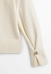 Pull tricoté couleur crème avec poignets côtelés et un bouton métallique décoratif sur la manche. Texture douce et coupe décontractée.