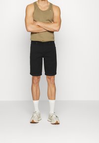 Homme portant un débardeur kaki, un short noir retroussé, des chaussettes blanches et des baskets beiges, se tenant avec les bras croisés devant un fond blanc.