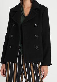 Žena v černém dvouřadém kabátu přes zelený top a vysokých pruhovaných kalhotách v černé, bílé a hnědé barvě.