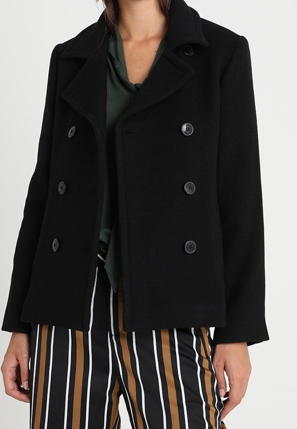 Žena v černém dvouřadém kabátu přes zelený top a vysokých pruhovaných kalhotách v černé, bílé a hnědé barvě.