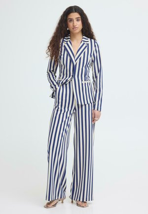 Femme aux longs cheveux bouclés portant un costume à rayures verticales bleues et blanches avec un pantalon à jambes larges et des sandales à talons, se tenant devant un fond uni.