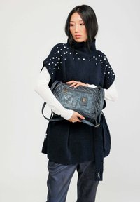 Bolso bandolera azul acolchado con una correa cruzada, que presenta acentos decorativos y un acabado texturizado, sostenido por un modelo con un vestido azul marino y mangas blancas.
