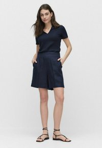 Laivansiniset poolopaita V-pääntiellä, yhdistettynä laivansinsisiin pellavashortseihin, joissa on sivutaskut, ja mustat nauhakiilakengät.