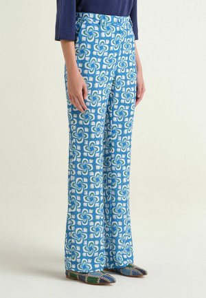 Donna che indossa pantaloni a gamba larga con motivo floreale blu e bianco, top blu navy e scarpe basse a punta con motivo patchwork.