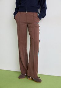 Braune Nadelstreifenhose mit ausgestelltem Bein, die blaue Akzente an den Seiten und ein gesticktes Logo am linken Oberschenkel aufweist.