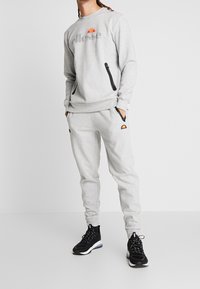 Sudadera gris con logo, bolsillos con cremallera lateral y pantalones de chándal gris a juego con acento de logo. Llevado con zapatillas deportivas negras.