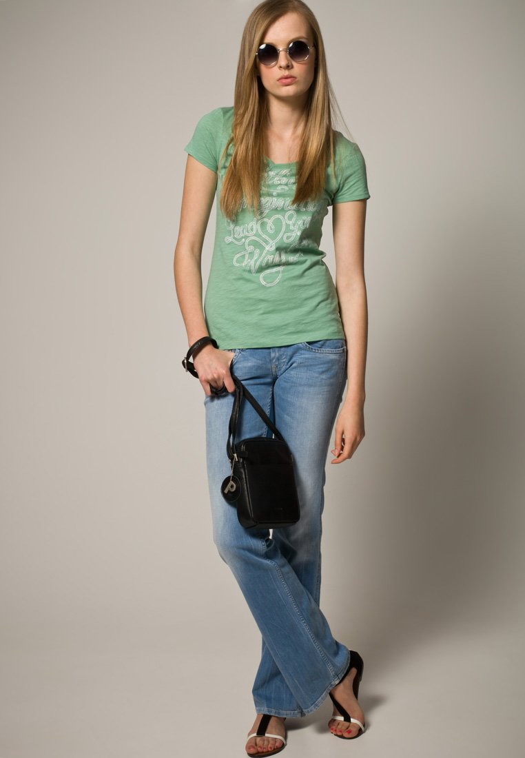 Junge Frau mit langen Haaren, die runde Sonnenbrillen trägt, ein grünes Grafik-T-Shirt, blaue Jeans und ein schwarzes Armband hat, während sie eine schwarze Handtasche hält.