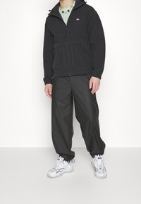 Svart vattentät jacka med dragkedja och huva, utrustad med en bröstficka och detaljerad sömnad. Kombinerad med löst sittande svarta byxor och vita sneakers.