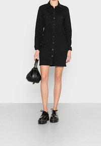 Musta farkkutunika, jossa on pitkät hihat, nappi-etumus, kaksi rintataskua ja naisellinen silhouette. Yhdistetty mustien platform-kenkien ja käsilaukun kanssa.