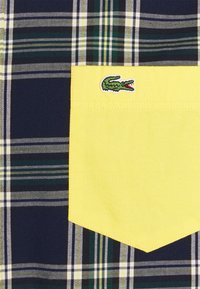 Osasto plaid-paitoja, jossa on tummansinisiä, vihreitä ja valkoisia raitoja. Keltainen tasku, jossa on vihreä krokotiililogo. Puuvillamateriaali, teksturoitu kangas.