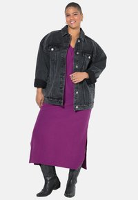 Schwarze Denim-Übergroßjacke mit Knopfverschlüssen, getragen über einem lila knielangen Kleid; kombiniert mit kniehohen schwarzen Cowboystiefeln.