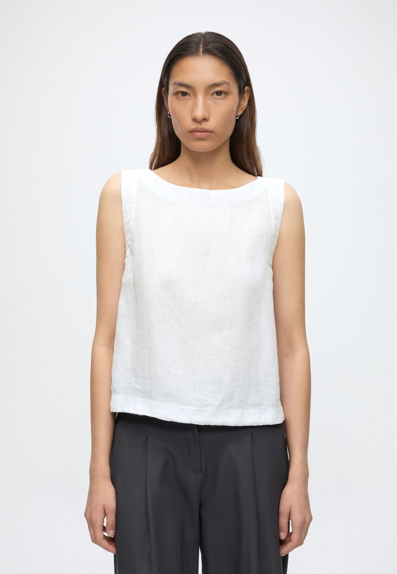 Jeune femme aux longs cheveux foncés, portant un haut blanc sans manches et un pantalon ample gris foncé, debout devant un fond clair uni.