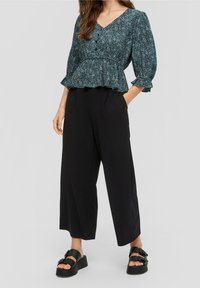 Pantaloni neri a gamba larga abbinati a un top peplo a maniche lunghe in teal con motivo, caratterizzato da scollatura a V e dettagli con bottoni. Sandali con plataforma neri.