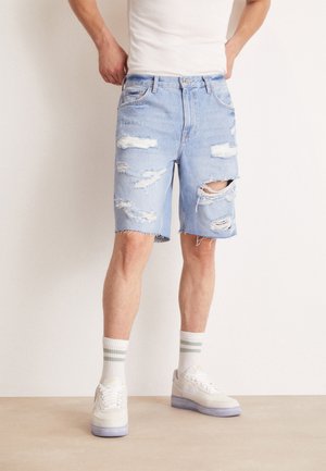 Shorts de mezclilla azul claro con detalles desgastados, múltiples rasgones, dobladillo deshilachado y diseño de cinco bolsillos. Combinados con zapatillas blancas y calcetines a rayas.