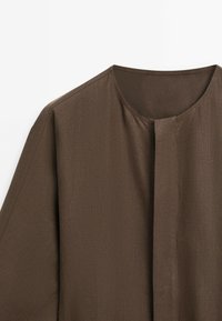 Vêtement marron texturé avec un col rond et une ouverture sur le devant, mettant en valeur la surface lisse du tissu et son design minimaliste.