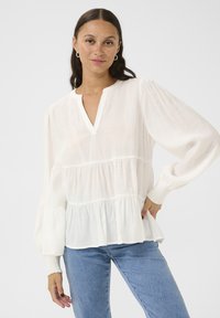 White, layered blouse with a V-neck and long puff sleeves. Features a textured fabric with elastic cuffs and a relaxed fit. Worn with blue jeans.