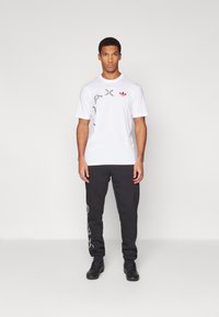 White short-sleeve t-shirt with graphic prints, paired with black sweatpants featuring side accents, and black sneakers.