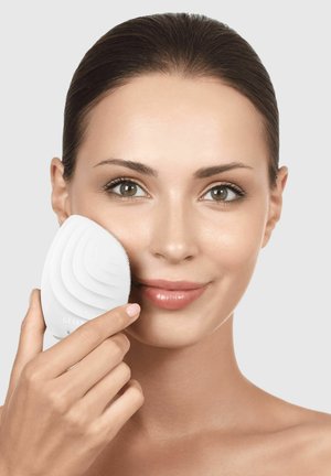 Woman with smooth skin holds a white, leaf-shaped facial cleansing brush to her cheek against a light background.