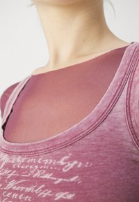 Topp med dyp U-halsdesign, laget av et mykt, teksturert stoff i dyp mauve. Overlayet har en semi-transparent, glatt overflate.