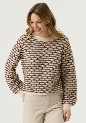 Woman smiling, wearing a beige sweater with brown scallop pattern and light beige pants, standing against a plain light background.