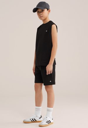 Jong persoon draagt een zwart mouwloos shirt en korte broek, grijze pet, witte sokken en witte Adidas-sneakers met zwarte strepen, staand.