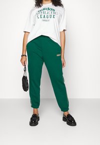 Vihreät collegehousut, joissa on valkoiset sivuraitat ja logopatchi, yhdistettynä valkoiseen ylisuureen t-paitaan, jossa on vihreät tekstit. Mustat kengät ja laukku.