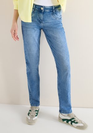 Persoon die lichtblauwe slim-fit jeans, witte en groene sneakers en een geel shirt draagt, staand tegen een effen lichte achtergrond.