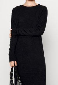 Černé svetrékové šaty s volným střihem, dlouhými rukávy, žebrovanými manžetami a širokým výstřihem. Měkká, texturovaná látka. Stylizováno s černou kabelkou.