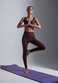Brown athleisure set with a cropped top and high-waisted leggings. Model stands on one leg on a purple yoga mat, hands in prayer position.