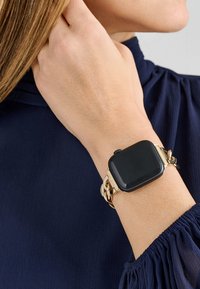 Smartwatch z kwadratową czarną tarczą i złotą bransoletą w stylu łańcucha. Bransoleta posiada błyszczące ogniwa i zapięcie. Noszona na nadgarstku o jasnym odcieniu skóry.