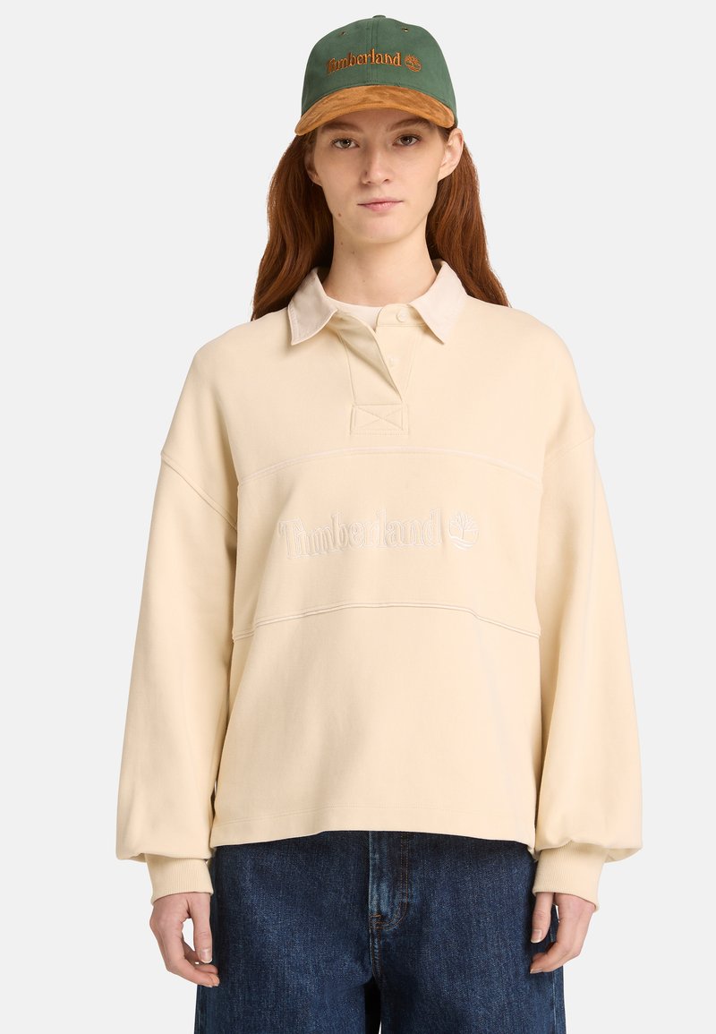 Sweat-shirt à col polo de couleur crème avec un logo brodé sur le devant. Associé à un short en denim bleu foncé et une casquette verte.