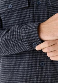 Dunkelgraue karierte Hemdjacke aus Wolle, mit blauen Knöpfen, einem knöpfbaren Bund und einem strukturierten Muster. Nahaufnahme des Ärmel-Details.