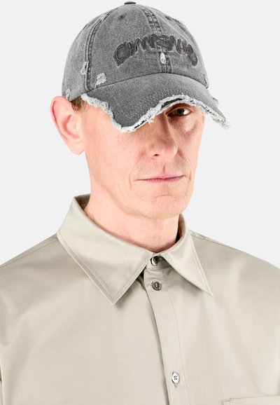 Gorra de béisbol gris desgastada con bordes deshilachados y texto bordado. Usada con una camisa de color beige claro con cuello que presenta detalles de botones.