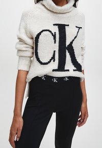 Biały sweter z dzianiny z czarnym logo "CK", z wysokim kołnierzem i ściągaczami na rękawach, w połączeniu z czarnymi spodniami z logo na pasku.