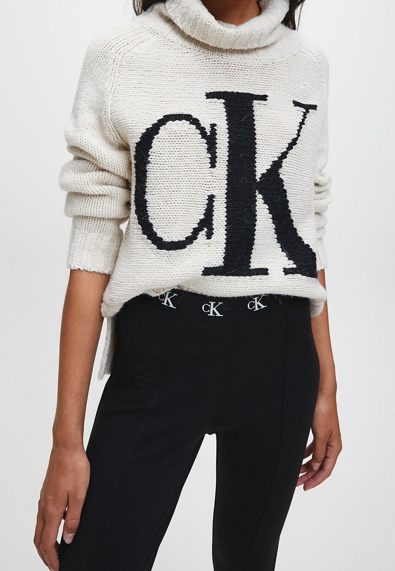 Biały sweter z dzianiny z czarnym logo "CK", z wysokim kołnierzem i ściągaczami na rękawach, w połączeniu z czarnymi spodniami z logo na pasku.