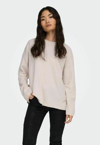 Sudadera de manga larga beige claro con un ajuste relajado y cuello redondeado, combinada con pantalones negros texturizados. Diseño sencillo, tejido suave.