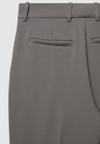 Pantalon gris sur mesure avec une texture lisse, deux poches arrière avec des coutures discrètes et des passants pour ceinture le long du haut.