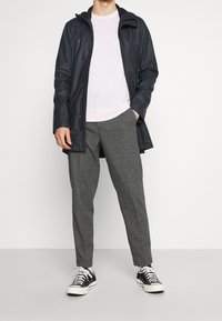 Svart vattentät jacka med huva, ljusrosa tröja under, grå strukturerade byxor och svarta sneakers med vita sulor, stående mot en vit bakgrund.