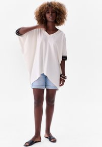 White oversized tunic with a V-neck, featuring black striped cuffs, paired with light denim shorts and black slide sandals.