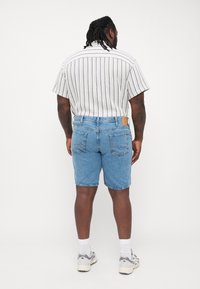 Pantalones cortos de mezclilla azul con dobladillo deshilachado, bolsillos traseros y una etiqueta de cuero color miel. Combinados con una camisa de rayas de manga corta y calcetines blancos.